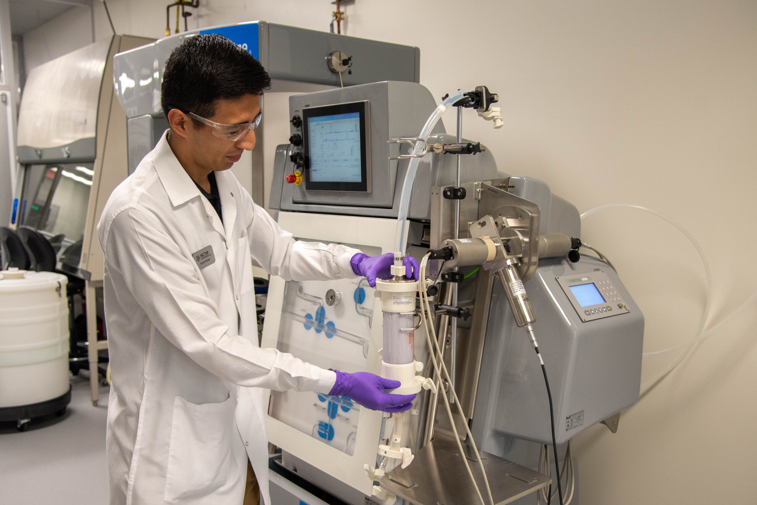 A scientist in a laboratory wearing a lab coat and safety goggles operates equipment while standing, focusing on the tools and instruments they are using.
