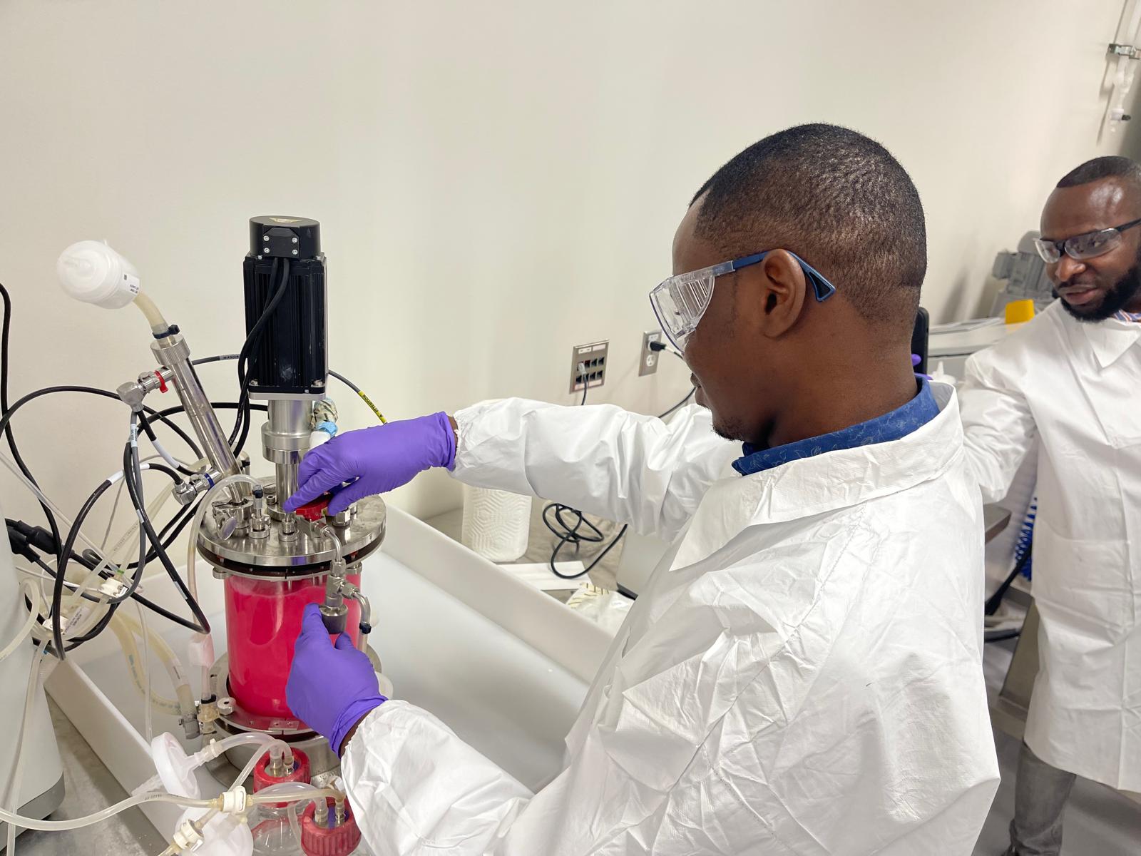 A scientist in a laboratory wearing a lab coat and safety goggles operates equipment while standing, focusing on the tools and instruments they are using.