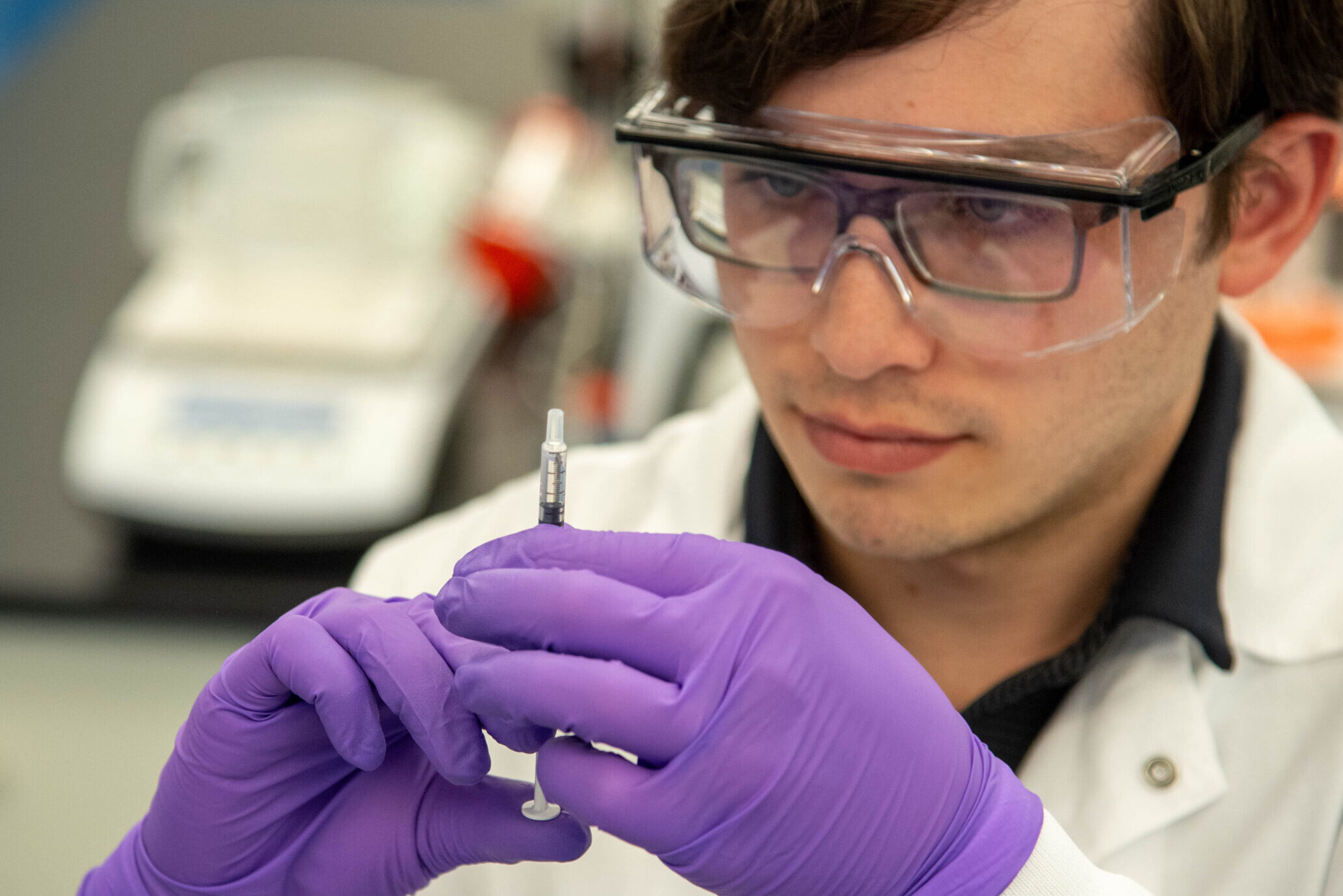 Scientist wearing protective goggles in a laboratory holding and closely examining a syringe, suggesting scientific or medical research